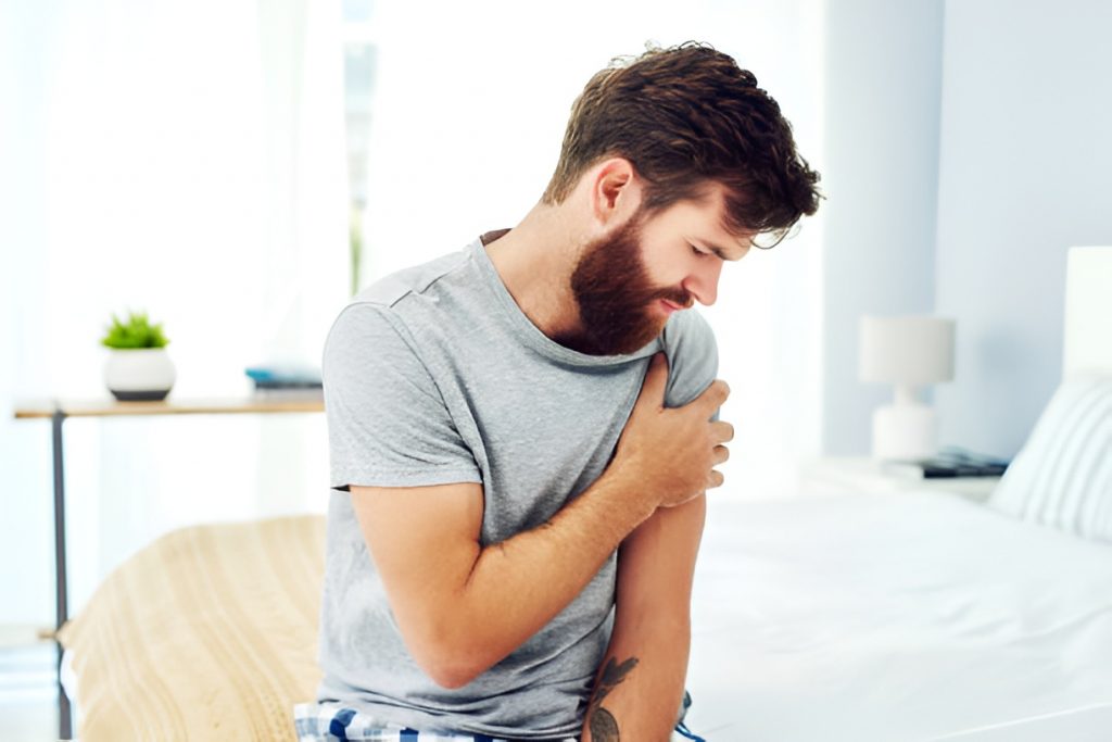 man sitting in bed, wincing in pain while squeezing his left arm