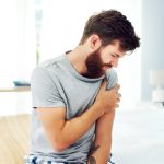 man sitting in bed, wincing in pain while squeezing his left arm